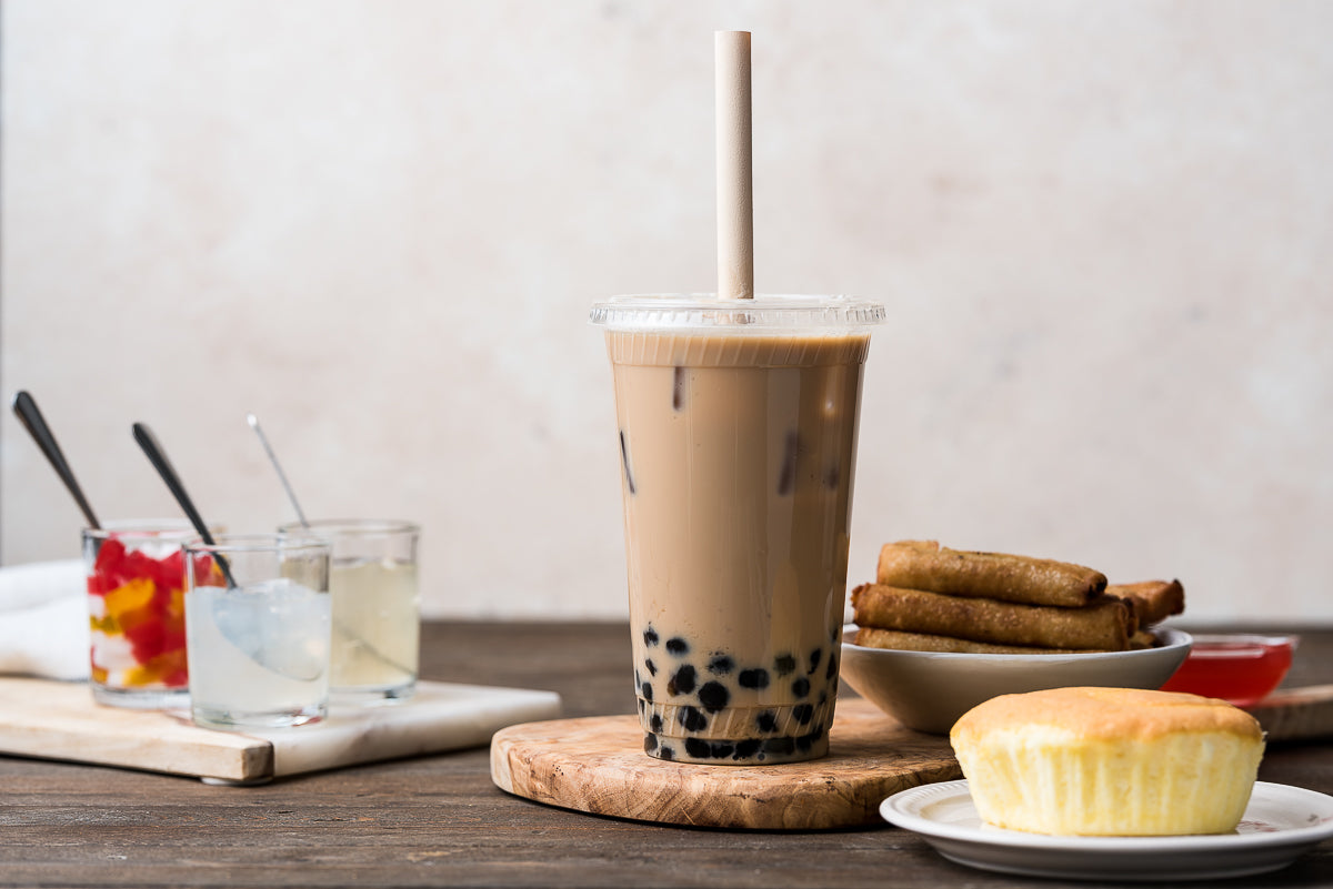 Boba straw in a bubble tea alongside a cupcake, spring roll, and three glasses with assorted beverages.